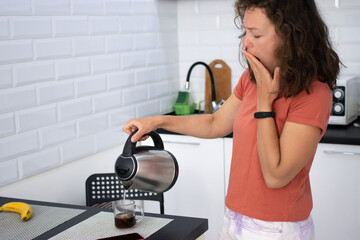 Sleepy young brunette woman yawning, covering her mouth with her palm, pouring boiled water from kettle, brewing strong coffee for waking up, standing in home kitchen.Lack of sleep, working all night