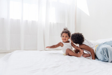 Obraz na płótnie Canvas Brother and sister African Americans play together on white bed in loft interior. Siblings having fun among the blue pillows in the morning. Boy kissing little girl on the cheek, caring for a neighbor