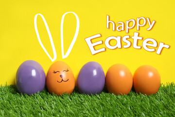 Happy Easter. One egg with drawn face and ears as bunny among others on green grass against yellow background