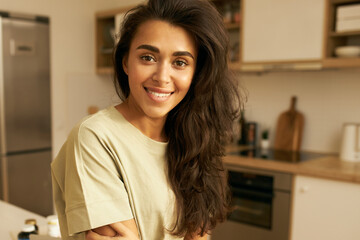 Fototapeta na wymiar People, relaxation, rest and leisure concept. Close up shot of adorable young Hispanic female in casual comfy t-shirt posing in kitchen interior, spending day at home, looking at camera with smile