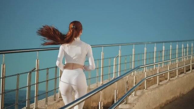 A back view of a young redhead woman wearing white sportsuit is running outside in the morning