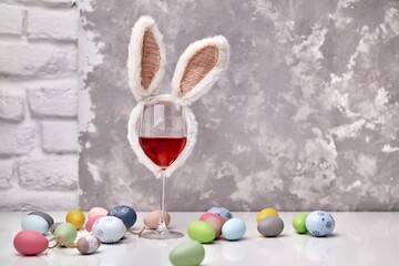 Glass of rose or red wine with bunny ears and Easter decorations, colorful eggs on white table, on bright background. Copy space for text.