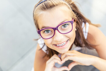 Young preteen girl in glasses wearing braces smiles at the camera showing heart shape with her hands
