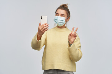 Positive, beautiful girl with blond hair gathered in bun. Wearing yellow sweater and protective face mask. Making selfie on a smartphone and showing peace sign. Stand isolated over white background
