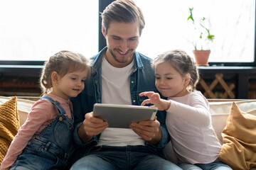 Beautiful young father, his cute little daughters are using a tablet and smiling, sitting on sofa...
