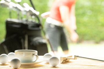 Golf balls and golf club and coffee cup on table on driving range with golfer in background