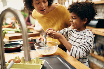 Happy black boy cracking egg while preparing food with mother in the kitchen.