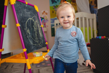 Naklejka premium A small blonde smiling girl at home in the game room near the blackboard with chalk in her hands