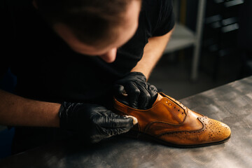 Top close-up view of professional shoemaker wearing black gloves polishing old light brown leather shoes. Concept of cobbler artisan repairing and restoration work in shoe repair shop. - Powered by Adobe