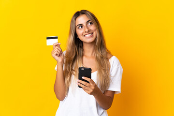 Naklejka premium Young hispanic woman over isolated yellow background buying with the mobile with a credit card while thinking