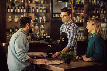 Happy waiter wearing face mask while serving coffee to a couple in cafe.