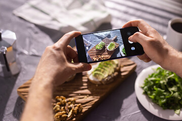Naklejka na ściany i meble Hands take pictures on smartphone of two beautiful healthy sour cream and avocado sandwiches lying on board on the table. Social media and food concept