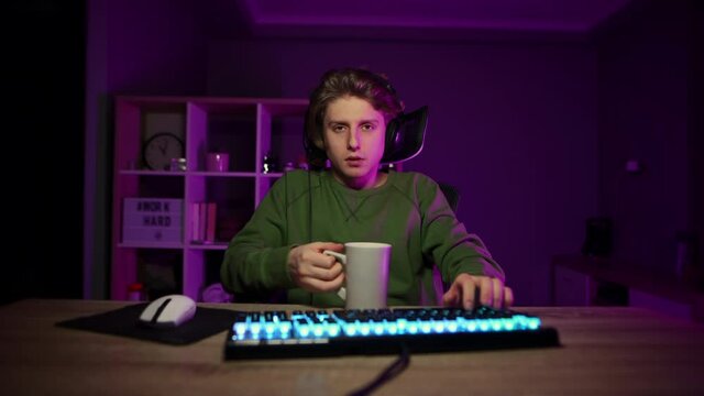 Tired young man sitting in a game chair in the room with a purple light, playing video games on the computer and drinking coffee from a cup. Night gaming.