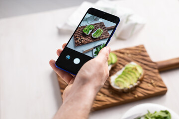 Hands take pictures on smartphone of two beautiful healthy sour cream and avocado sandwiches lying on board on the table. Social media and food concept