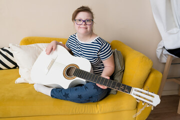 Young teenage girl with Down syndrome learning to play the guitar sitting on sofa at home. Musical...