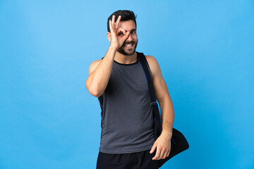 Young sport man with sport bag isolated on blue background showing ok sign with fingers