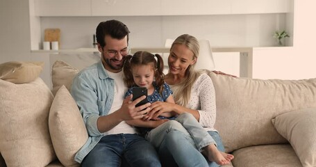 Happy attractive couple and small daughter spend weekend together using smartphone, family relaxing on couch in cozy living room teach kid showing new cool funny application. Modern tech usage concept
