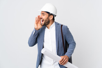 Young architect man with helmet and holding blueprints isolated on white background shouting with mouth wide open to the side