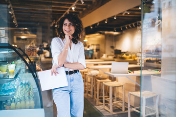 Happy female manager with menu in hand standing in doorway of local cafeteria and smiling, pretty...