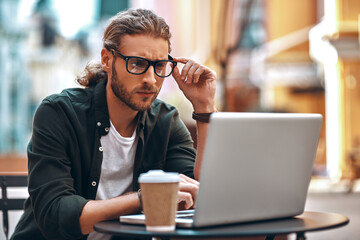 Busy young man in casual clothing working using computer and drinking coffee while sitting in cafe