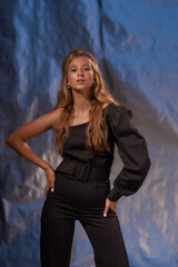 Young elegant blonde woman with long wavy hair and fashionable make-up with blue eye arrows wearing black overall, standing and posing against blue mettalic background