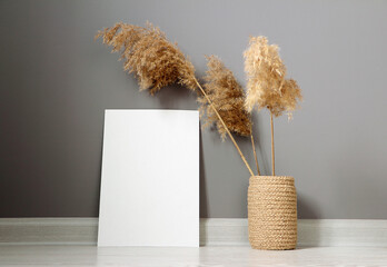 Empty mockup poster. Blank vertical picture on background gray wall. Dry reed in a knitted jute vase. Minimal interior design. Modern style.