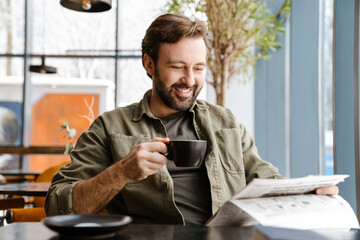 Unshaven smiling man drinking coffee while reading newspaper in cafe - Powered by Adobe