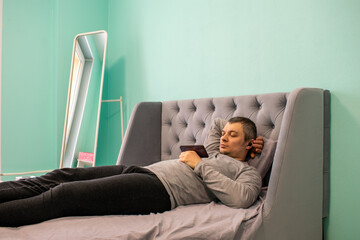 The young man, freelancer lies in the bed and watches the video or plays on the phone. Light green wallpapers, grey furniture and white floor mirror.