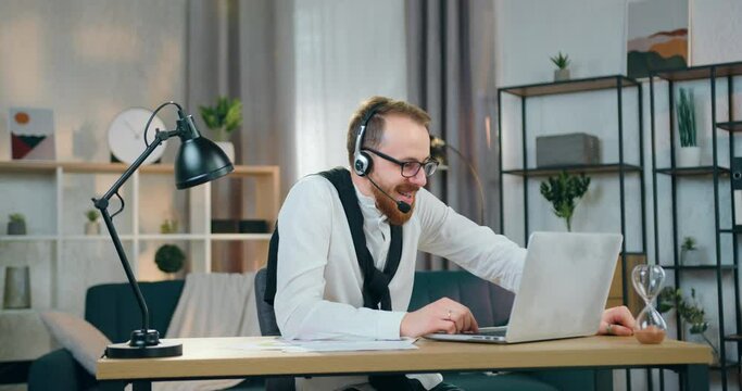 Handsome smiling in good mood young bearded guy in headset sitting in front of computer at home office and discussing details of charts report with colleague via video call