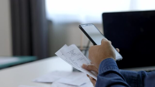 A young man is using a smartphone to scan the bill to calculate his income and expenses. In his hand there were bills to pay a lot in debt.
