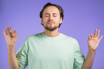 Calm man in basic t-shirt relaxing, meditating. He calms down, breathes deeply with mudra om on violet studio background. Yoga concept.