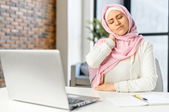 Tired Middle-eastern female worker kneading her neck, muscle pain after long day of work, sit at office desk, massaging neck, feeling ache, having spasm symptoms, feel unwell from sedentary lifestyle