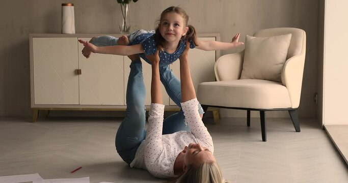 Mom lying on floor in light living room raises cute little daughter on outstretched hands, family performs acroyoga exercises, lead an active sporty lifestyle spend time together at modern cozy home