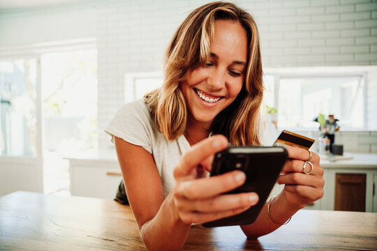 Caucsasian female student leaning against counter smiling making online payment holding credit card and cellula device
