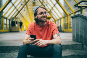 Stylish hipster smiling with smartphone while sitting on steps