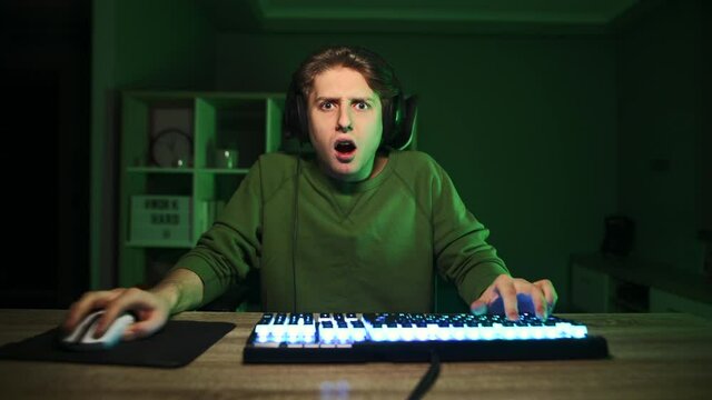 Surprised young male gamer in headset playing video games on computer at home and looking at camera with shocked face. Emotional gaming