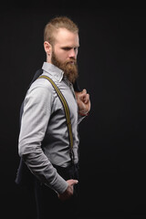 Portrait of a brutal attractive bearded Caucasian man in a shirt with suspenders and a jacket on his shoulder. Studio photography