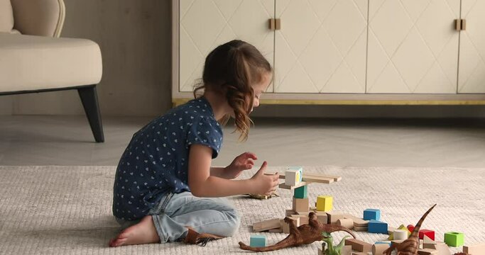 Small 4s girl sit on warm floor on carpet build with wooden cubes alone in living room, having fun enjoy playtime with playthings and dinosaurs toys set. Children development, leisure activity concept