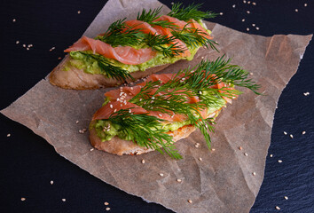 two slices of crispy baguette greased with avocado and decorated with smoked salmon fillets and sprigs of fresh dill, lie on parchment paper