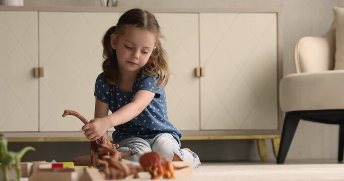 Small 4s adorable kid girl with ponytails having fun alone at home play dinosaur toys set sitting in light warm modern playroom. Imagination creativity development, hobby and interest of child concept