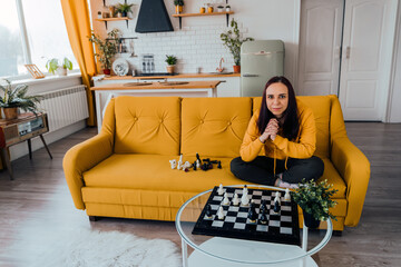 Young woman sitting on yellow sofa and playing chess in room. Female playing in logical board game with herself.