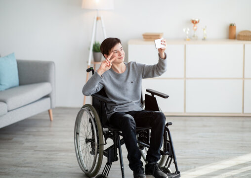 Happy disabled teenager in wheelchair taking selfie on smartphone, smiling at camera indoors