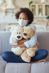 Cute mixed race teen girl in casual clothes wearing protective face mask, looking at camera, hugging her toys while sitting on a sofa at home