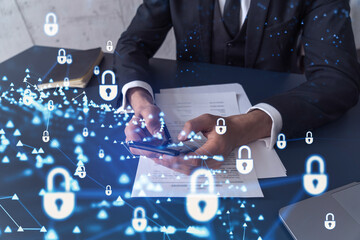 A businessman in formal wear reading the contract to prevent probability of risks in cyber security and checking details in the phone. Padlock Hologram icons over the working desk.