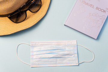 A Protective medical face mask, travel book, hat and glasses  on a blue background. Travel concept during the Coronavirus, Covid-19 pandemic, flatlay