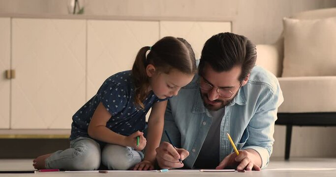 Caring young father teach cute little daughter drawing, family sit on floor in modern living room holding pencils create paintings in sketchbook together. Happy fatherhood, home hobby and fun concept