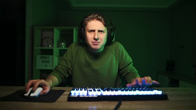 Upset gamer in a headset cries after a loss, sitting at night in a bedroom with a purple light. An emotional gamer in despair cries after losing.