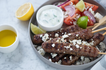 Close-up of bbq beef kofta kebabs with greek tzatziki, feta cheese and vegetable salad, studio shot on a grey marble surface