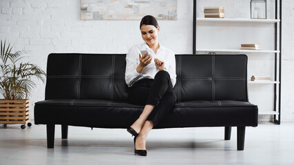 joyful businesswoman sitting on couch and messaging on mobile phone