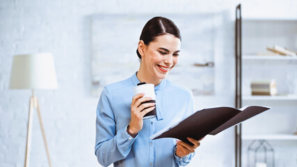 Obraz premium cheerful businesswoman holding coffee to go and folder with documents in office